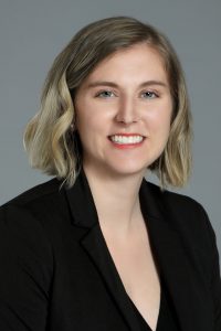 Alishia smiles in a black suit coat in front of a blue background for a portrait photo